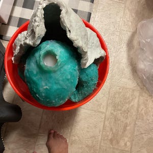 Op de afbeelding: Een rode emmer met een turquoise, onregelmatig gevormd object met een witte substantie erin. Een grijs, gekreukeld materiaal bedekt het object gedeeltelijk. De emmer staat op een betegelde vloer.