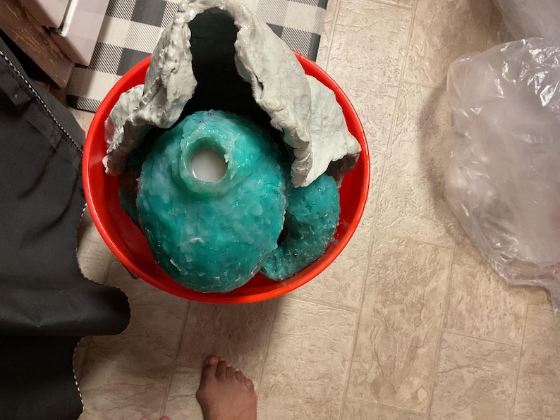 Op de afbeelding: Een rode emmer met een turquoise, onregelmatig gevormd object met een witte substantie erin. Een grijs, gekreukeld materiaal bedekt het object gedeeltelijk. De emmer staat op een betegelde vloer.