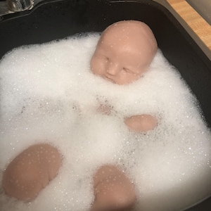 Op de afbeelding: Een poppenhoofd en ledematen ondergedompeld in zeepwater in een zwarte gootsteen. De poppenonderdelen hebben een huidskleur en het water is gevuld met witte bellen. De gootsteen staat tegen een lichtgekleurd aanrecht.