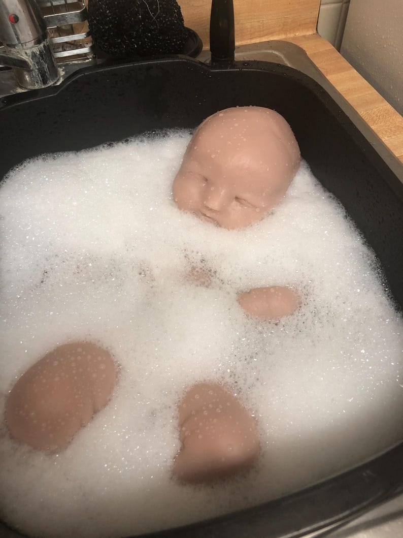 Op de afbeelding: Een poppenhoofd en ledematen ondergedompeld in zeepwater in een zwarte gootsteen. De poppenonderdelen hebben een huidskleur en het water is gevuld met witte bellen. De gootsteen staat tegen een lichtgekleurd aanrecht.