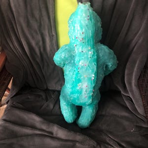 Op de afbeelding: Een turquoise, handgemaakte sculptuur van een wezen met een lange staart en kleine ledematen. De sculptuur is versierd met kleine, kleurrijke spikkels. Het figuur zit op een donkergrijze deken.