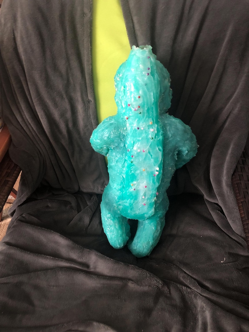 Op de afbeelding: Een turquoise, handgemaakte sculptuur van een wezen met een lange staart en kleine ledematen. De sculptuur is versierd met kleine, kleurrijke spikkels. Het figuur zit op een donkergrijze deken.