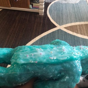 Op de afbeelding: Een grote, turquoise, amorfe sculptuur met een glanzende afwerking. De sculptuur is versierd met kleine, kleurrijke sprinkles. Het object wordt in de hand van een persoon gehouden.