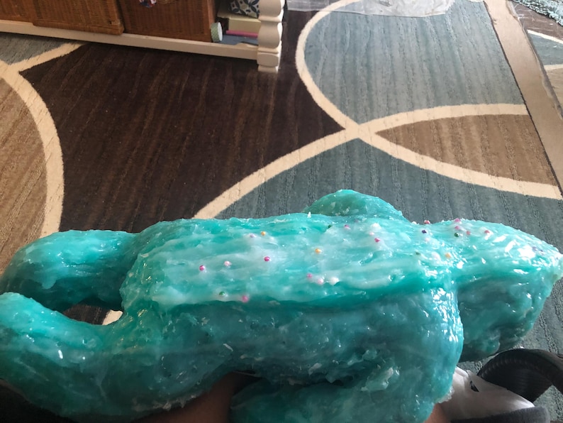 Op de afbeelding: Een grote, turquoise, amorfe sculptuur met een glanzende afwerking. De sculptuur is versierd met kleine, kleurrijke sprinkles. Het object wordt in de hand van een persoon gehouden.