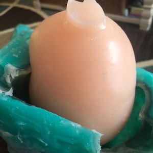 Op de afbeelding: Een licht perzikvormig, afgerond object met een gebroken, doorschijnende bovenkant, genesteld in een turquoise, getextureerde mal. De mal heeft een golvende, organische vorm. Het object lijkt een knutselartikel te zijn.