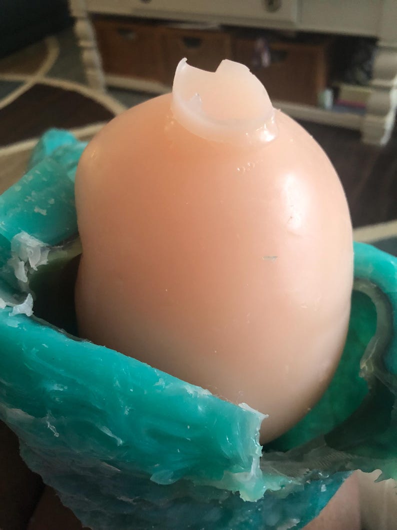 Op de afbeelding: Een licht perzikvormig, afgerond object met een gebroken, doorschijnende bovenkant, genesteld in een turquoise, getextureerde mal. De mal heeft een golvende, organische vorm. Het object lijkt een knutselartikel te zijn.