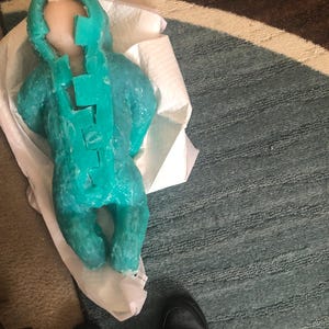 Op de afbeelding: Een turquoise wassen sculptuur van een baby, liggend op een witte papieren handdoek. De sculptuur heeft een gedetailleerd ontwerp, met een hoofd en lichaam. De baby ligt op een blauw en grijs tapijt.