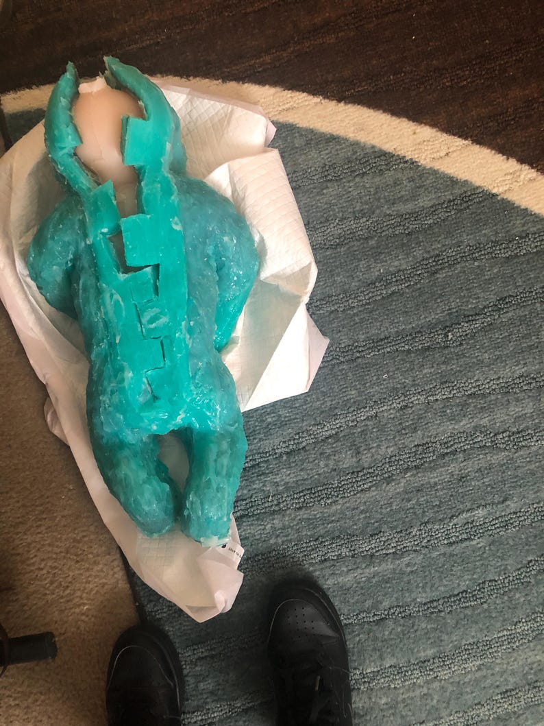 Op de afbeelding: Een turquoise wassen sculptuur van een baby, liggend op een witte papieren handdoek. De sculptuur heeft een gedetailleerd ontwerp, met een hoofd en lichaam. De baby ligt op een blauw en grijs tapijt.