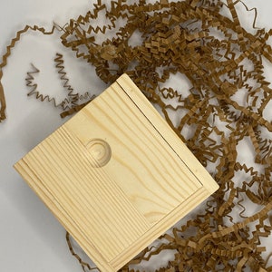 Op de afbeelding: Een kleine, vierkante houten doos met een scharnierend deksel. De doos is gemaakt van licht hout en heeft een natuurlijke afwerking. Het deksel heeft een kleine, ronde knop in het midden. De doos staat op een bed van bruin gekreukt papier.