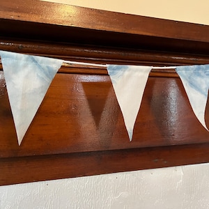 Może przedstawiać: Girlanda trójkątnych chorągiewek z tkaniny z niebiesko-białym wzorem tie-dye. Chorągiewki są nawleczone na biały sznurek i zawieszone na tle ciemnego drewna.