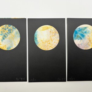 May include: Three abstract art prints featuring circular designs in shades of yellow, blue, and white on a black background. Each print has a unique color pattern and is numbered. The prints are displayed side by side.