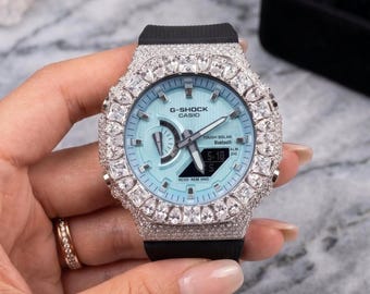 Może przedstawiać: Zegarek G-Shock Casio z jasnoniebieską tarczą i czarnym paskiem. Zegarek jest inkrustowany przezroczystymi kryształami wokół ramki. Zegarek jest trzymany w dłoni ze złotym pierścionkiem.