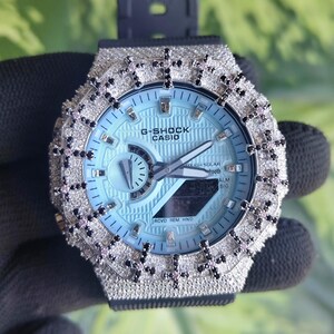 Può includere: Un orologio G-Shock Casio con quadrante azzurro e cinturino nero. L'orologio è incrostato di pietre color argento e dettagli neri attorno alla lunetta. Il quadrante mostra il logo G-Shock Casio.