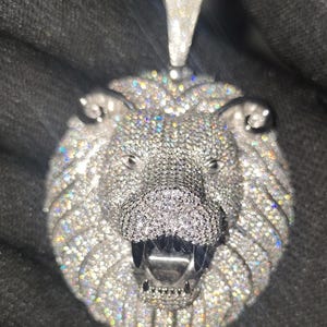 May include: A silver-toned lion pendant encrusted with numerous small, sparkling gemstones. The pendant features a detailed lion's head with an open mouth, visible teeth, and a bail for a chain. The gemstones reflect light, creating a dazzling effect.