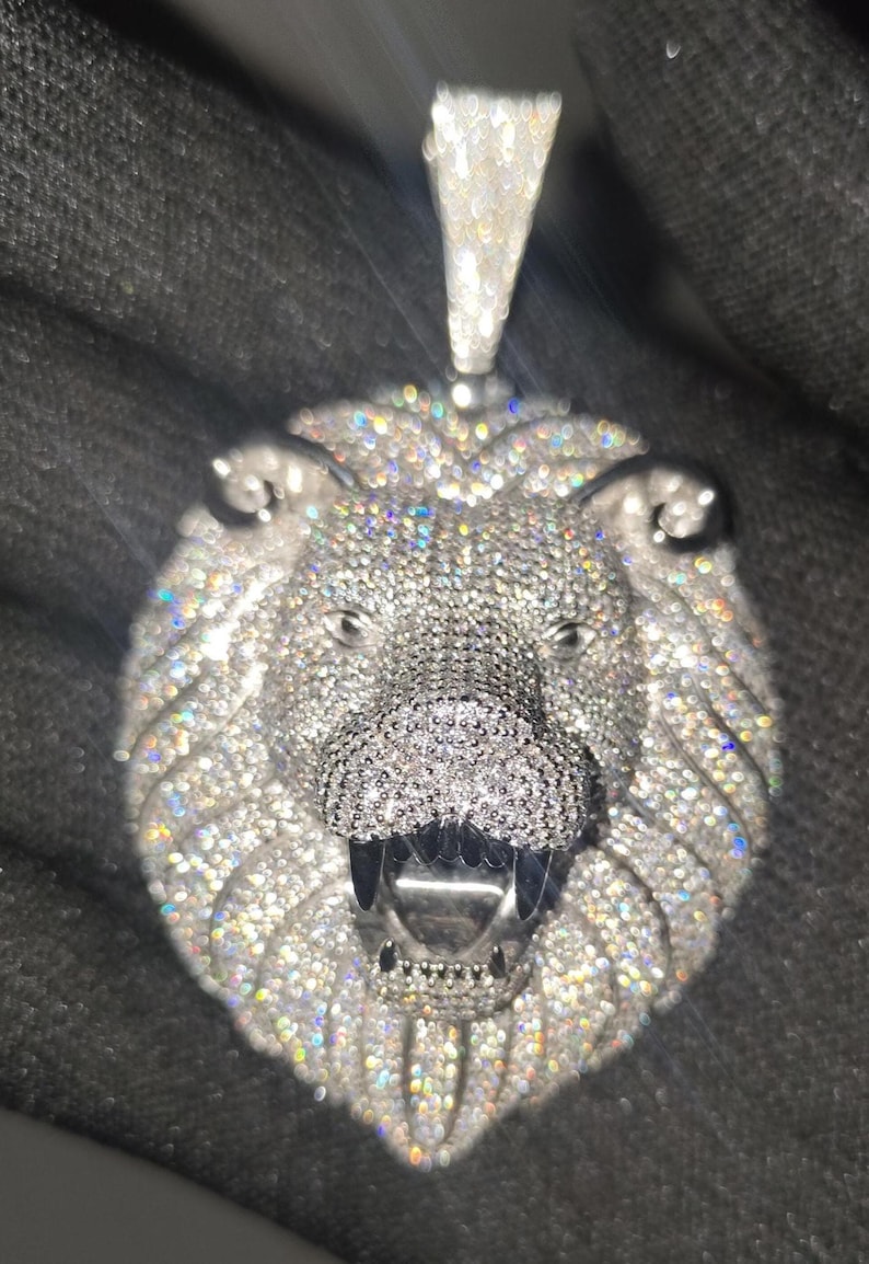 May include: A silver-toned lion pendant encrusted with numerous small, sparkling gemstones. The pendant features a detailed lion's head with an open mouth, visible teeth, and a bail for a chain. The gemstones reflect light, creating a dazzling effect.