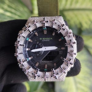 Può includere: Un orologio G-Shock Casio con quadrante analogico-digitale nero, lancette bianche e una lunetta incastonata con numerose piccole pietre chiare, arricchita da motivi a croce con pietre nere e rosa. Include un cinturino in tessuto verde oliva.