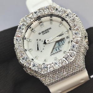 May include: A white G-Shock Casio watch featuring an octagonal bezel and case fully encrusted with clear, sparkling stones. The watch has a white analogue-digital dial with silver hands, markers, and the text G-SHOCK CASIO, 20BAR, and MUTE ALM SIG.