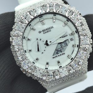 May include: White G-Shock Casio watch with a diamond-encrusted bezel and strap. The watch face is white with black hands and a digital display. The watch shows the time and date. The watch is set against a dark background.