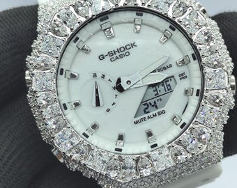 May include: White G-Shock Casio watch with a diamond-encrusted bezel and strap. The watch face is white with black hands and a digital display. The watch shows the time and date. The watch is set against a dark background.