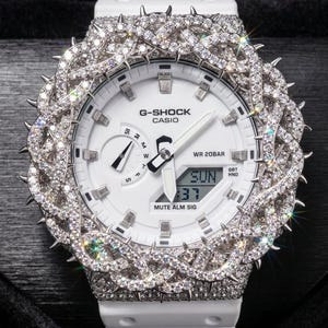 May include: White G-Shock watch with a diamond-encrusted bezel and strap. The watch face displays the day of the week, time, and the G-Shock and Casio brand names. The watch has a digital display and is water resistant to 20 bar.