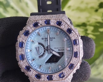 May include: A G-Shock watch with a light blue face, surrounded by a silver bezel with clear and blue stones. The watch has a black band and displays the time. The watch face has the words "G-Shock" and "Bluetooth".