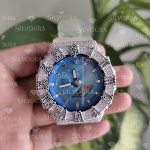 May include: A G-Shock wristwatch featuring a bright blue analog-digital dial and a bezel encrusted with numerous clear stones. The bezel includes black stone Roman numeral hour markers and a small green dollar sign emblem at the twelve o'clock position.
