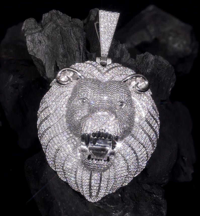 May include: A silver lion pendant encrusted with sparkling gemstones. The lion's head is detailed with an open mouth, visible teeth, and a textured mane. The pendant includes a diamond-studded bail for a chain.
