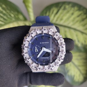 Può includere: Un orologio G-Shock con cinturino blu navy e quadrante blu scuro. La lunetta dell'orologio è incastonata con pietre preziose chiare e nere. L'orologio visualizza l'ora e altre funzioni. L'orologio è tenuto da una mano guantata.