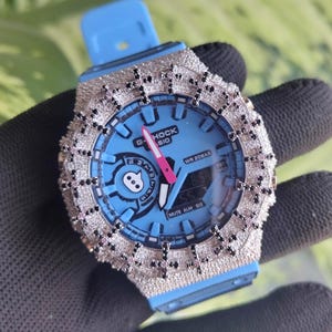 Può includere: Un orologio G-Shock blu con una lunetta tempestata di diamanti e un cinturino blu abbinato. Il quadrante dell'orologio è azzurro con una lancetta dei secondi rosa e la scritta "G-Shock". L'orologio è tenuto da una mano guantata.