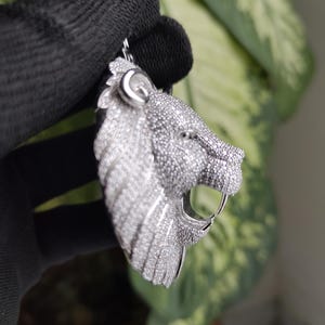 May include: A silver pendant in the shape of a lion's head, encrusted with small, sparkling stones. The lion's mouth is open, revealing a single fang. The pendant is held by a black gloved hand.