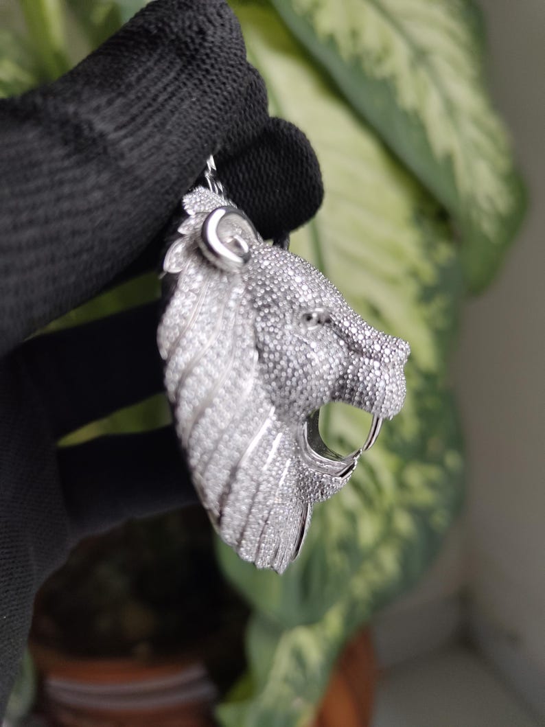 May include: A silver pendant in the shape of a lion's head, encrusted with small, sparkling stones. The lion's mouth is open, revealing a single fang. The pendant is held by a black gloved hand.