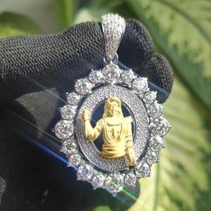 May include: A gold and silver pendant featuring a central gold figure within a diamond-studded halo. The pendant has a diamond-encrusted bail and a circular design, with a detailed figure in the center. The pendant is held against a dark background.