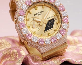 May include: A gold G-Shock Casio watch featuring a bezel encrusted with alternating pink and white heart-shaped stones and small pavé crystals. The watch face displays the text G-Shock Casio, WR20BAR, and digital time. Text reads: CUSTOMIZED TO PERFECTION. FEATURING HEART-SHAPED DIAMOND BEZEL and BESPOKE HEARTS & GOLD COLLECTION.