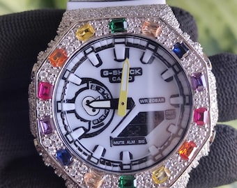 May include: White G-Shock watch with a silver bezel encrusted with small, clear crystals and colorful gemstones. The watch face is white with black accents and the text "G-SHOCK". The watch band is white.