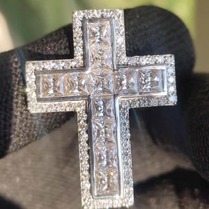 May include: A silver cross pendant with a square-cut gemstone center and a border of smaller, round gemstones. The cross is held against a dark background, highlighting the sparkle of the jewelry.