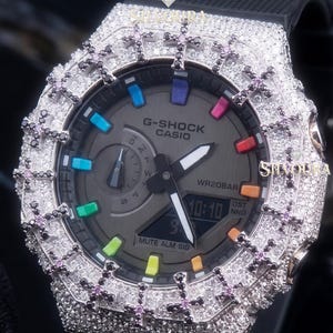 Può includere: Un orologio G-Shock Casio con quadrante nero, indici multicolori e display digitale. La lunetta ottagonale è incastonata con pietre chiare e decorata con motivi a croce in pietre nere e rosa. È dotato di un cinturino nero.