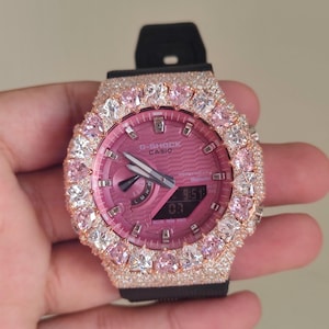 Könnte beinhalten: Eine G-Shock Casio-Uhr mit rosa Zifferblatt und schwarzem Armband. Die Uhr ist mit rosa und klaren Edelsteinen um die Lünette besetzt. Das Zifferblatt zeigt die Uhrzeit 9:51 an.