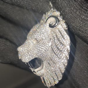 May include: A silver-colored lion pendant encrusted with numerous small, sparkling stones. The lion's head is detailed with an open mouth and a textured mane. The pendant is designed to be worn as jewelry.