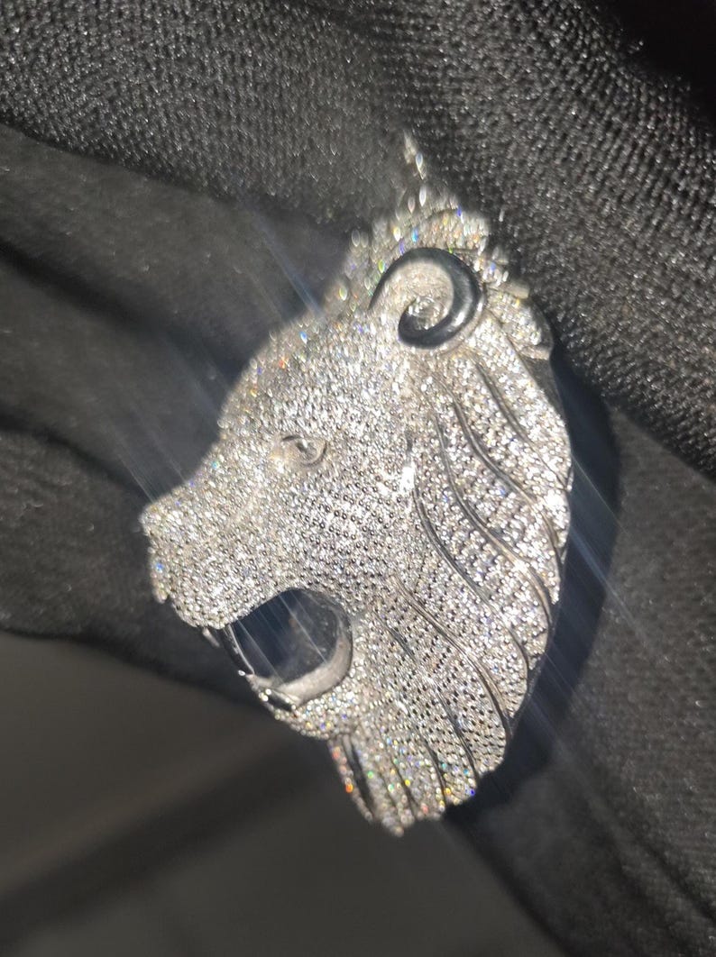 May include: A silver-colored lion pendant encrusted with numerous small, sparkling stones. The lion's head is detailed with an open mouth and a textured mane. The pendant is designed to be worn as jewelry.