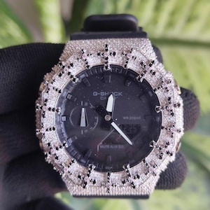 Può includere: Un orologio G-Shock nero con una lunetta argentata incastonata con piccole pietre scintillanti. Il quadrante dell'orologio è nero con lancette e indicatori bianchi. Il cinturino dell'orologio è nero.