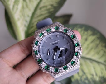 Może przedstawiać: Zegarek G-Shock Casio z szarą tarczą i szarym paskiem. Ramka zegarka jest inkrustowana małymi białymi kamieniami i większymi zielonymi kamieniami. Zegarek jest trzymany w dłoni.
