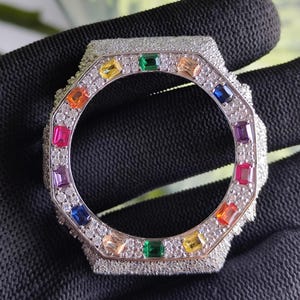 Puede incluir: Un bisel de reloj plateado con forma octogonal. El bisel está incrustado con pequeñas piedras preciosas transparentes alrededor del borde exterior y presenta un anillo de piedras preciosas de colores en varios colores, incluyendo rojo, naranja, amarillo, verde y azul.