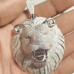 May include: A silver-colored lion pendant with a detailed design. The lion's head is covered in small, sparkling stones, with an open mouth revealing sharp teeth. The pendant has a loop at the top for a chain.