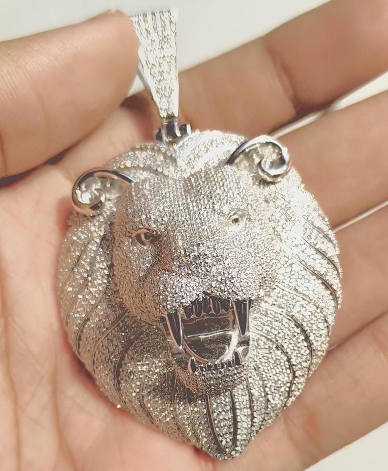 May include: A silver-colored lion pendant with a detailed design. The lion's head is covered in small, sparkling stones, with an open mouth revealing sharp teeth. The pendant has a loop at the top for a chain.