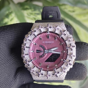 Può includere: Un orologio G-Shock Casio con quadrante rosa e lunetta argentata incastonata con piccole pietre scintillanti. L'orologio ha un cinturino in gomma nero. Il quadrante mostra l'ora. L'orologio è tenuto da una mano guantata.