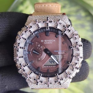 Può includere: Un orologio G-Shock Casio con quadrante marrone e lunetta incastonata di pietre chiare, caratterizzato da accenti a forma di croce neri e rosa. Il quadrante mostra il testo G-Shock, Casio, Tough Solar, Bluetooth e Mute Alm Sig.
