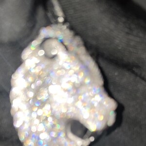 May include: A silver pendant in the shape of a lion's head, encrusted with numerous sparkling, clear gemstones. The pendant has a bail at the top for a chain. The gemstones reflect a rainbow of colors.