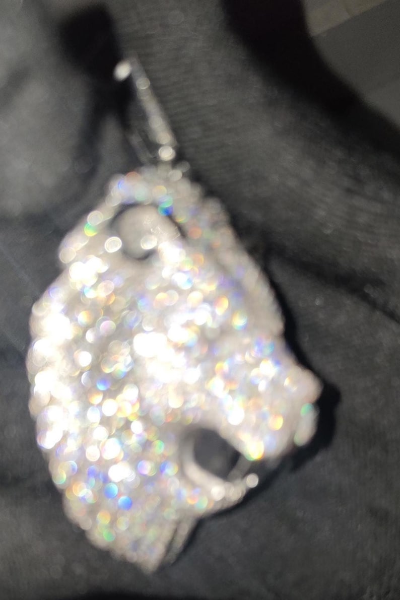 May include: A silver pendant in the shape of a lion's head, encrusted with numerous sparkling, clear gemstones. The pendant has a bail at the top for a chain. The gemstones reflect a rainbow of colors.