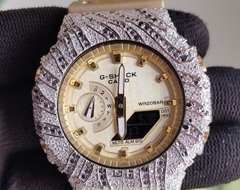 Może przedstawiać: Złoty zegarek na rękę z wysadzaną diamentami ramką i paskiem. Tarcza zegarka jest złota z czarnymi detalami i napisem "G-SHOCK CA 0". Zegarek posiada wyświetlacz cyfrowy i jest trzymany przez czarną rękawicę.