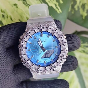 Può includere: Un orologio da polso argentato con quadrante azzurro e display digitale che mostra l'ora. L'orologio è incastonato con piccole pietre scintillanti attorno alla lunetta. Il cinturino è grigio chiaro.